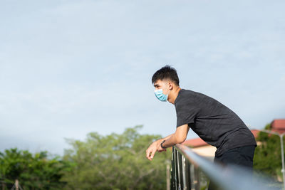 Side view of young man looking away standing by railing against sky