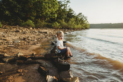 Side view of baby boy sitting on rock at lakeshore against sky during sunset