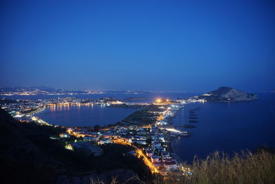Illuminated city by sea against clear blue sky at night