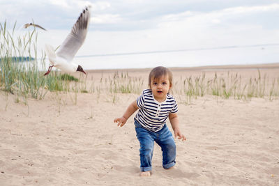 Baby boy in striped sailor t-shirt running on the sandy beach with seagulls near the sea in summer