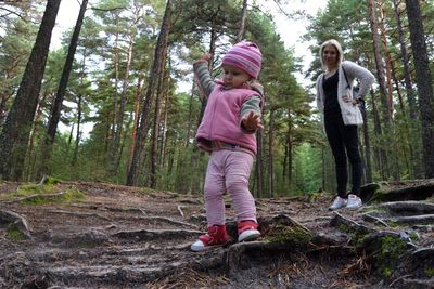 Woman looking at daughter standing on roots against trees in forest