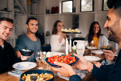 Young man holding bowl of strawberries while sitting with friends for lunch party