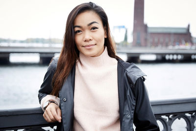 Portrait of smiling young woman standing by railing on bridge in city