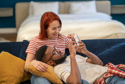 Young diverse couple using smartphone choose hotel for vacation while resting on sofa at home