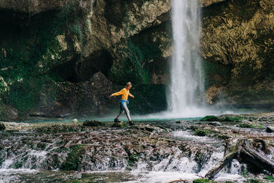 A young active woman in a yellow longsleeve walks across a stream in a nature reserve.