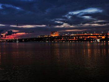 Illuminated city by sea against sky at sunset