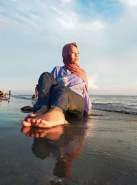 Young woman sitting at beach against sky