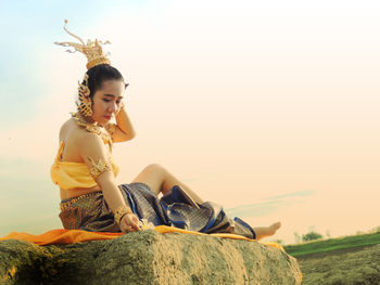 Full length of woman in traditional clothing sitting on rock against sky during sunset