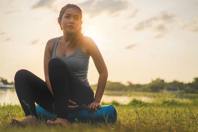 Young woman sitting on grass at field against sky during sunset