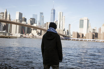 Rear view of man standing by river and buildings against sky