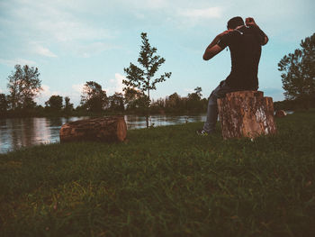Rear view of man sitting on tree stump by lake during sunset