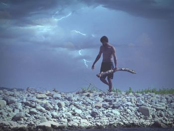 Full length of shirtless man carrying log on rocks against cloudy sky