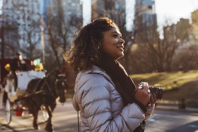 Side view of smiling woman holding digital camera on city street