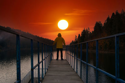 Rear view of man walking on footbridge against sky during sunset