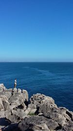 Man standing on cliff by sea against clear blue sky