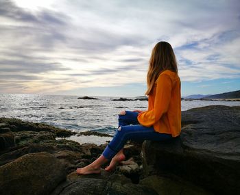 Side view of woman sitting on rock at beach against cloudy sky