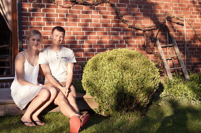 Full length portrait of smiling couple sitting in yard against brick wall