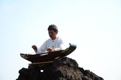 Low angle view of man sitting on rock against clear sky