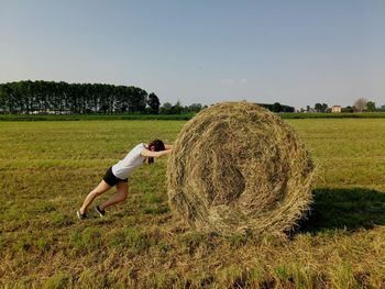 Side view of woman rolling hay bale on field against sky
