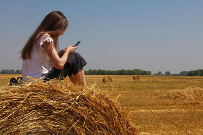 Side view of woman using mobile phone while sitting on hay bale at farm against clear sky