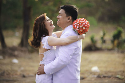 Side view of bride holding bouquet embracing groom on field