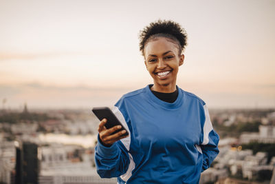 Portrait of smiling sportswoman holding smart phone during sunset