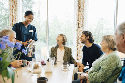 Smiling female nurse serving drink to senior woman sitting with friends and grandson at dining table in nursing home