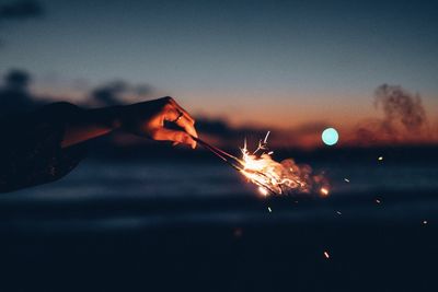 Cropped hand holding burning sparkler at sunset against sky