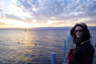 Portrait of smiling woman standing on pier over sea against cloudy sky during sunset