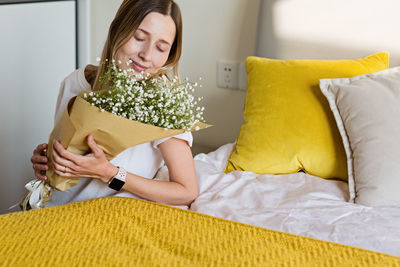 Beautiful woman holding bouquet sitting by bed at home