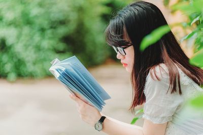 Side view of woman reading book while sitting against plants