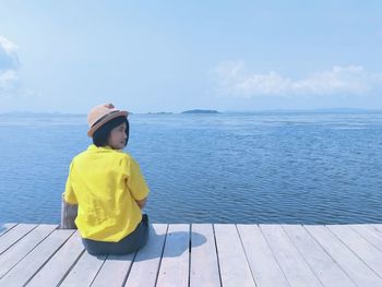 Rear view of boy sitting on sea shore against sky