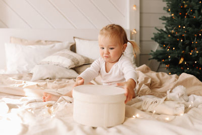 Funny funny little girl baby opens christmas gift box sitting on bed at home on holiday