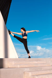 Full body of sporty female in activewear split and stretching leg on wall during outdoor training against blue sky on summer day