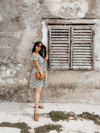 Front view of young woman in polka dot dress posing in front of weathered wall and old window.