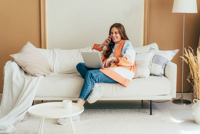 Young confident busy red-haired woman in a cardigan sitting on the sofa calling on the phone.