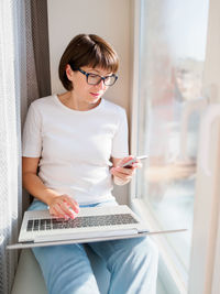 Woman using phone while sitting with laptop on sill