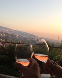 Close-up of hand holding glass of cityscape against sky during sunset