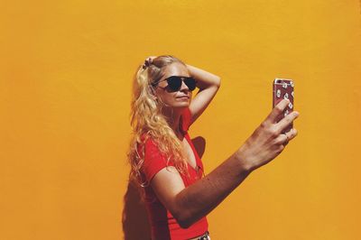 Woman taking selfie while standing against orange wall during sunny day