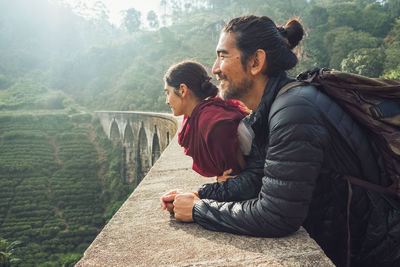 Side view of cheerful ethnic man and woman in activewear with backpacks standing contemplating views on aged stone nine arch bridge among green hills in sri lanka