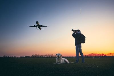 Mid adult man photographing airplane while standing by dog on field against sky during sunset