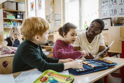 Smiling teacher assisting boy and girl with digital tablet sitting at desk in classroom