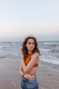 Dreamy young female with long hair looking away while standing on sandy beach near waving sea