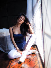 Full length of ballet dancer sitting by curtain at home