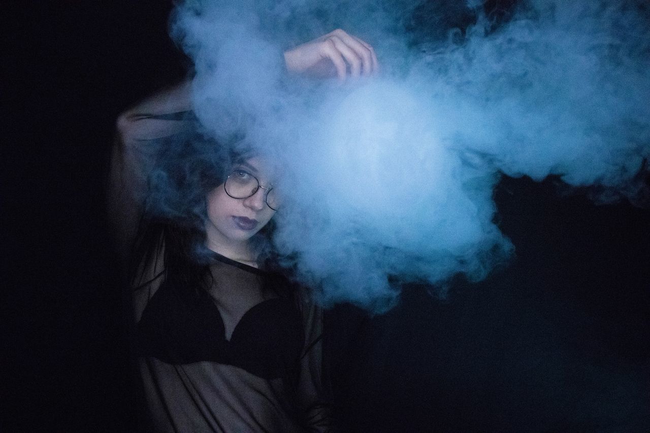 one person, smoke - physical structure, indoors, young adult, real people, front view, lifestyles, leisure activity, night, standing, casual clothing, motion, smoking issues, waist up, three quarter length, bad habit, black background, dark, hairstyle