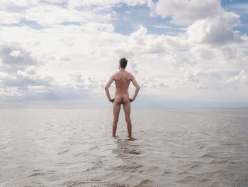 Rear view of naked man standing with arms akimbo in sea against sky