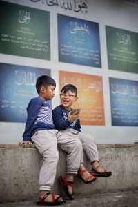 Male friends using phone while sitting against text on wall