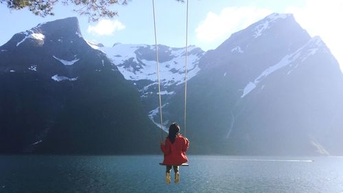 Rear view of woman sitting on swing by lake against mountain