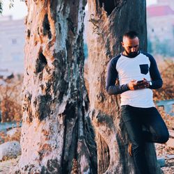 Young man using phone while standing against tree trunk