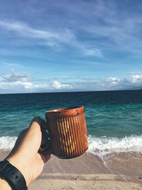 Cropped hand holding coffee cup at beach against sky